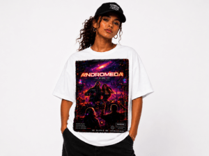 Woman modeling a white oversized T-shirt with a neon space-poster graphic reading 'ANDROMEDA' and a cosmic scene in red and purple hues, cap on head, hands in pockets.