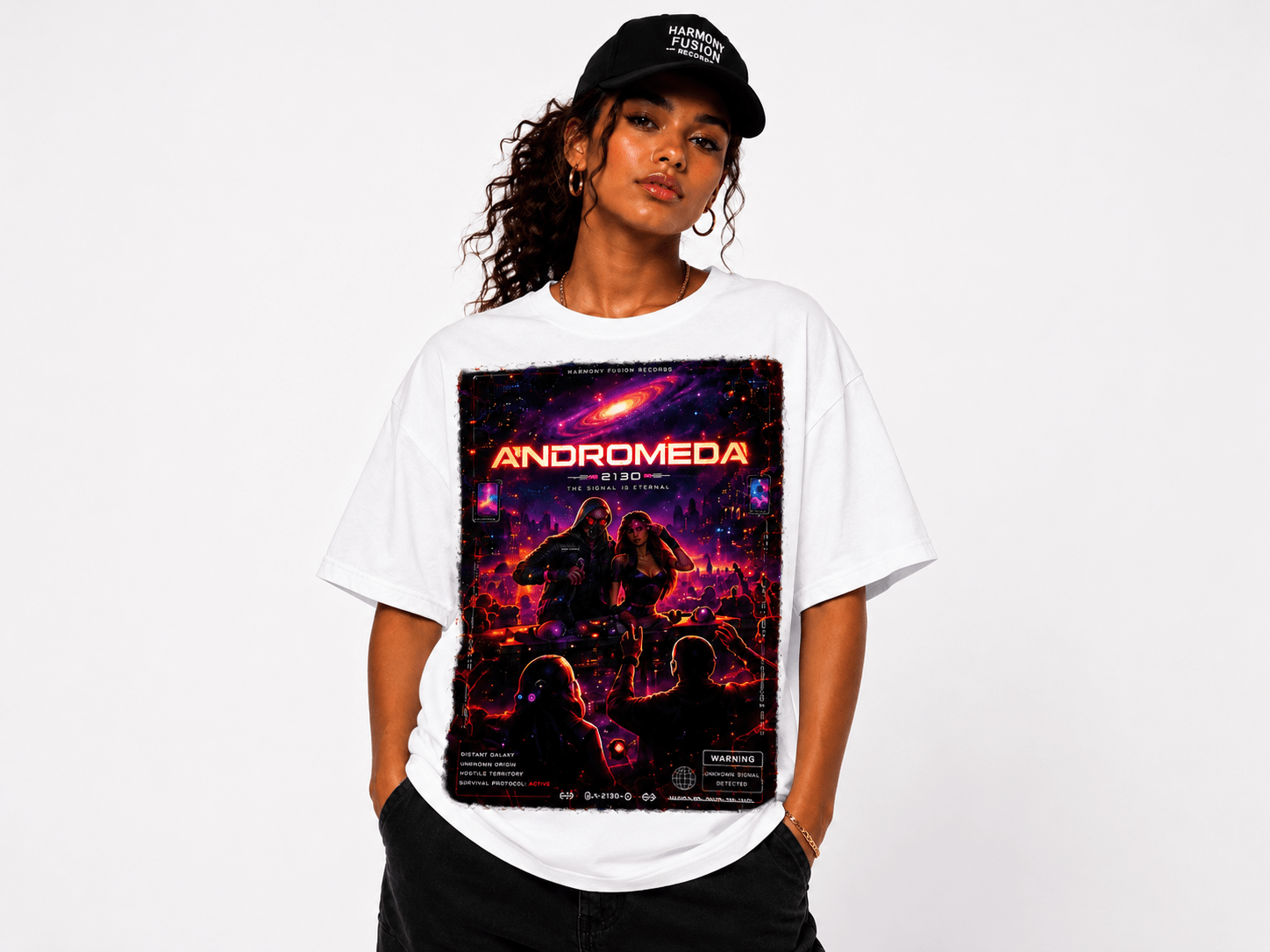 Woman modeling a white oversized T-shirt with a neon space-poster graphic reading 'ANDROMEDA' and a cosmic scene in red and purple hues, cap on head, hands in pockets.