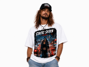 Man wearing a white graphic T-shirt with a bold city-at-night design reading 'STATIC SKIES'; he has long hair, a black cap, and layered bracelets.