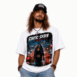 Man wearing a white graphic T-shirt with a bold city-at-night design reading 'STATIC SKIES'; he has long hair, a black cap, and layered bracelets.